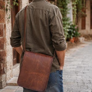 Könnte beinhalten: Eine braune Umhängetasche aus Leder mit rechteckiger Klappe und einem langen, verstellbaren Schulterriemen. Die Tasche hat eine glatte, abgenutzte Textur und wird quer über dem Körper getragen, wobei sie auf der Hüfte aufliegt. Das Design ist schlicht.