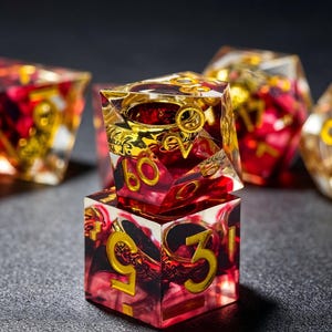 May include: A set of polyhedral dice made of clear resin with deep red swirling interiors and metallic gold numbering. The dice feature intricate gold-colored filigree patterns embedded within the transparent material, with a cube-shaped die stacked beneath a d20 die.