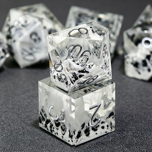 May include: A set of polyhedral dice made of clear resin with an internal black and white mountain landscape design. The dice feature silver-colored numbers etched into the surfaces. One die is stacked on top of another, showing clear facets.