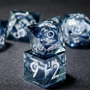 Op de afbeelding: Een set doorzichtige blauwe polyhedrale dobbelstenen van hars met zilverkleurige cijfers. De dobbelstenen hebben een wervelend glittereffect aan de binnenkant. Een tienzijdige dobbelsteen rust op een zeszijdige, met andere wazige dobbelstenen op de achtergrond.