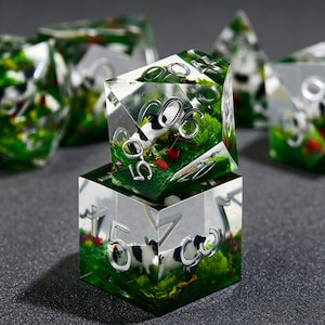 May include: A set of polyhedral dice made of clear resin, each containing miniature black and white cow figurines, green grass, and small red flowers. The dice feature silver-colored numbers embedded within the transparent material.