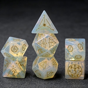 Puede incluir: Un juego de dados poliédricos hechos de material blanco lechoso translúcido con un brillo iridiscente. Cada dado presenta números grabados de color dorado y símbolos celestiales intrincados, incluyendo estrellas y lunas crecientes.