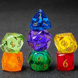 Può includere: Una collezione di dadi poliedrici a più facce in vari colori traslucidi, tra cui blu, viola, verde, giallo, arancione e rosso. Ogni dado presenta numeri incisi riempiti con inchiostro color oro, impilati in tre colonne verticali distinte.