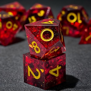 May include: A set of polyhedral dice made from translucent deep red resin with embedded gold-coloured flakes and glitter. The dice feature engraved numbers filled with gold paint. One ten-sided die is balanced on top of a six-sided die.
