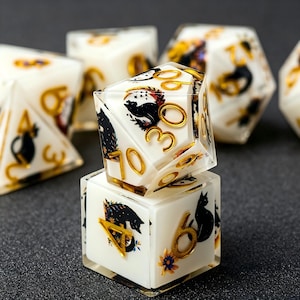 Puede incluir: Un juego de dados poliédricos hechos de resina blanca opaca con siluetas de gatos negros incrustadas, motivos florales y números de color dorado. Los dados presentan bordes afilados y una capa exterior transparente.