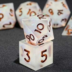 May include: A set of polyhedral dice made from translucent, iridescent white resin with copper-colored numbers. The dice feature sharp edges and include a ten-sided die with numbers 10, 90, and 04, and a six-sided die showing the numbers 3 and 5.