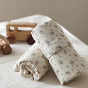 Könnte beinhalten: Zwei gerollte Stoffstücke in Off-White mit einem zarten Blumenmuster in gedeckten Blau- und Brauntönen. Der Stoff ist mit einem passenden Band befestigt und zeigt eine weiche, strukturierte Optik mit kleinen, sich wiederholenden botanischen Motiven.