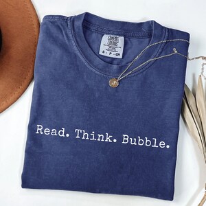 May include: A navy blue short-sleeve t-shirt with a crew neckline and a faded wash finish. The shirt features the white text "Read. Think. Bubble." printed across the chest. A small gold-toned circular pendant necklace rests on the collar area.