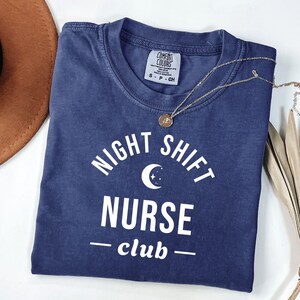 Peut inclure: Un t-shirt bleu marine à manches courtes avec une impression graphique blanche indiquant NIGHT SHIFT NURSE club, accompagnée d'une icône de croissant de lune et d'étoiles. Le t-shirt présente un col rond et une étiquette Comfort Colors.