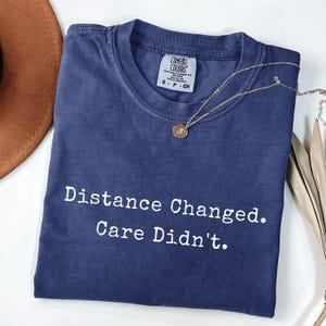 May include: A navy blue short-sleeve t-shirt with a crew neckline and a white printed graphic that reads Distance Changed. Care Didn't. The shirt features a Comfort Colors brand label on the inside collar indicating a size small.