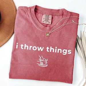 May include: A dusty rose short-sleeve t-shirt with a crew neckline. The shirt features the white text "i throw things" printed above a white line-art graphic of hands shaping clay on a pottery wheel. A small tag inside the collar reads Comfort Colors.