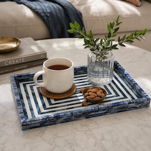 May include: A rectangular tray featuring a blue and white geometric chevron pattern. On the tray sits a white ceramic mug on a round woven coaster, a small brass bowl filled with almonds, and a clear ribbed glass vase containing green leafy stems.