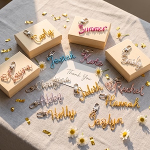 Puede incluir: Una colección de llaveros personalizados de alambre en varios colores, incluyendo oro, plata, rosa y azul, cada uno con un cierre de mosquetón. Varios llaveros están sobre cajas de regalo beige, junto a una tarjeta que dice Thank You.