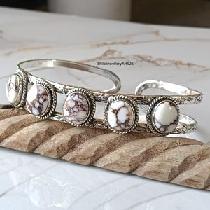 May include: A silver cuff bracelet featuring five oval-shaped white stones with brown veining. The metal band has a twisted rope border around each stone and engraved floral patterns along the sides. The text ShilaJewelleryArt925 is visible.