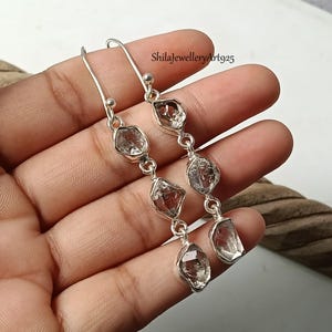 Op de afbeelding: Een paar lange oorbellen met zilveren haken en elk drie bungelende, heldere, ruw geslepen kristallen stenen. De metalen zettingen zijn van gepolijst zilver. De tekst ShilaJewelleryArt925 is zichtbaar in het bovenste deel van de afbeelding.