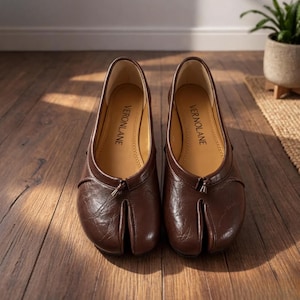 May include: A pair of dark brown split-toe shoes with a textured finish and a small tassel detail at the center front. The interior soles are tan and feature the brand name VERONLANE printed in black text on the heel area of each shoe.