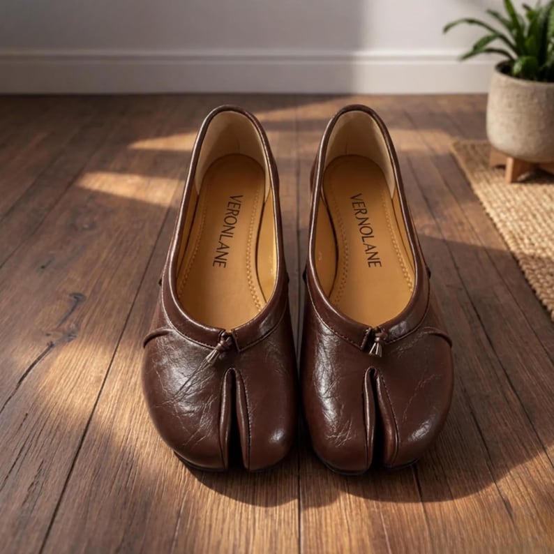 May include: A pair of dark brown split-toe shoes with a textured finish and a small tassel detail at the center front. The interior soles are tan and feature the brand name VERONLANE printed in black text on the heel area of each shoe.