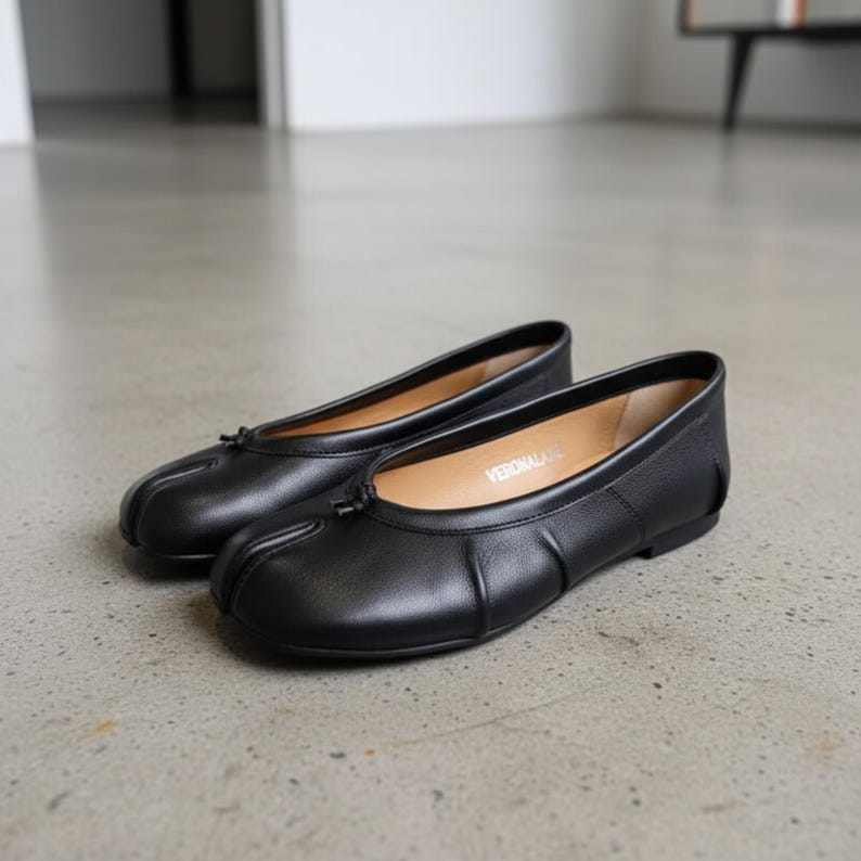 May include: A pair of black leather ballet flats featuring a split-toe design, a small decorative bow at the center of the vamp, and a low heel. The interior insole is tan and features the brand name VERONALATE printed in white lettering.