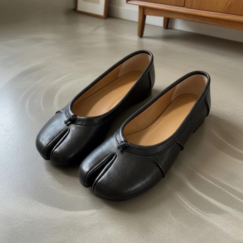 May include: A pair of black leather ballet flats featuring a split-toe design, a low heel, and a small decorative knot detail at the center of the vamp. The interior of the shoes is lined with a light tan material, providing a contrast to the black exterior.