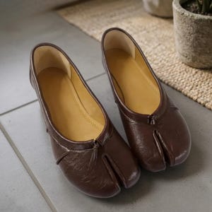 Op de afbeelding: Een paar platte schoenen van donkerbruin leer met een opvallend gespleten teenontwerp aan de voorzijde, een gladde beige voering en een laag profiel. De schoenen hebben een subtiel gestructureerde afwerking aan de buitenkant en een klein knoopdetail.