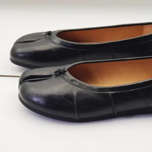 May include: A pair of black leather ballet flats featuring a distinct split-toe design at the front. Each shoe has a smooth, polished finish, a low-profile silhouette, and a small decorative knot detail centered at the top of the toe box.