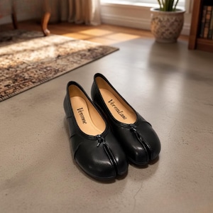 May include: A pair of black split-toe flats with a smooth finish and a small central knot detail at the vamp. The interior insoles are light beige and feature the brand names Veronne and Vermalane printed in black script on the respective footbeds.