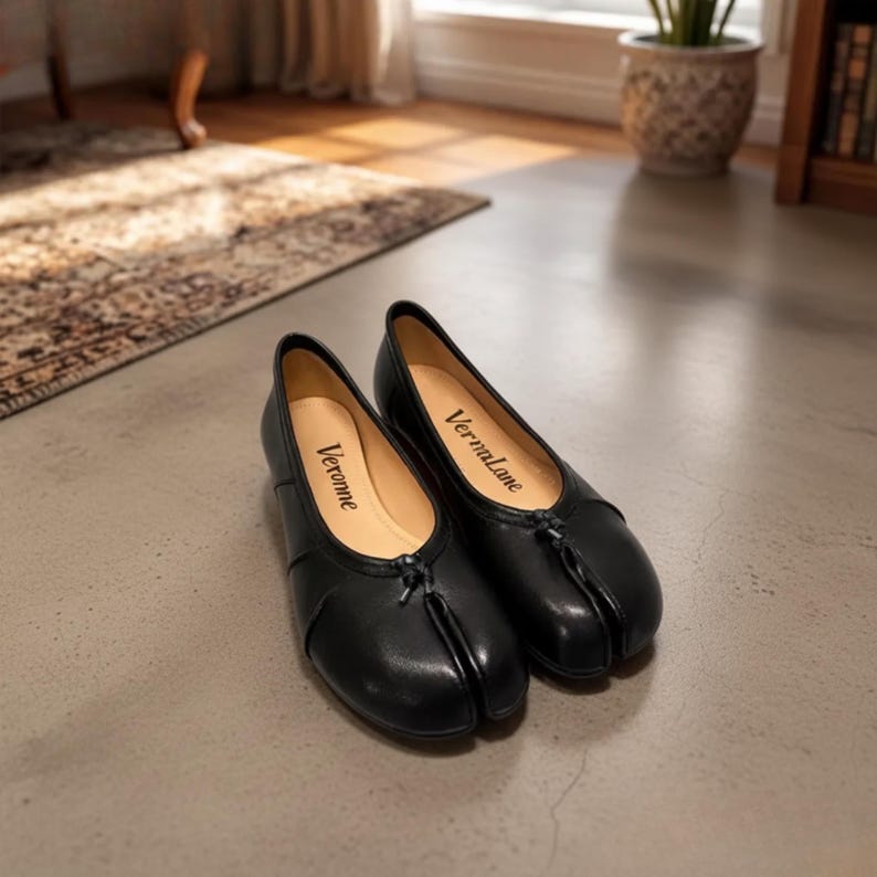 May include: A pair of black split-toe flats with a smooth finish and a small central knot detail at the vamp. The interior insoles are light beige and feature the brand names Veronne and Vermalane printed in black script on the respective footbeds.