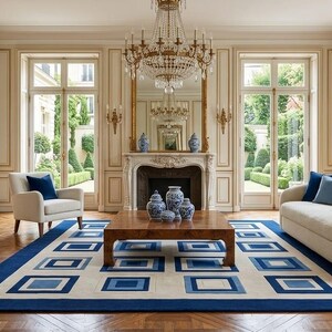 May include: A living room featuring a cream-colored sofa with blue pillows, a cream armchair with a blue pillow, and a wooden coffee table topped with blue and white ceramic vases. A large rug with a blue and cream geometric square pattern.
