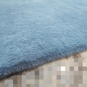 Op de afbeelding: Een dik, pluche vloerkleed in een effen stoffige blauwe kleur. Het kleed heeft een dicht, zacht getextureerd pooloppervlak met een strakke, rechte rand die de dikte van het materiaal onthult. De vezels zijn uniform en consistent.