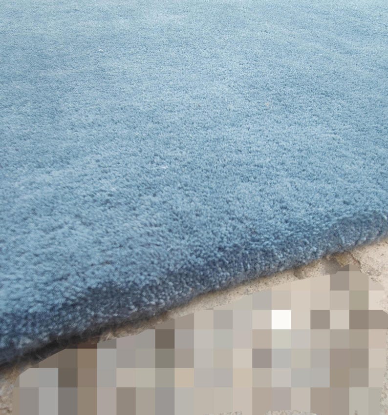 Op de afbeelding: Een dik, pluche vloerkleed in een effen stoffige blauwe kleur. Het kleed heeft een dicht, zacht getextureerd pooloppervlak met een strakke, rechte rand die de dikte van het materiaal onthult. De vezels zijn uniform en consistent.