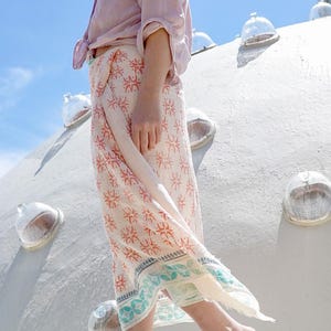 May include: A person wearing a light purple button-down shirt and a patterned skirt. The skirt features a repeating orange and white design with a teal and white border. The person is walking outside on a sunny day.