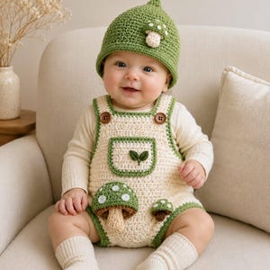 May include: A cream-colored crochet romper with green trim, featuring two brown wooden buttons, a front pocket with a leaf appliqué, and two mushroom appliqués. The outfit includes a matching green pointed crochet hat with a small mushroom detail.