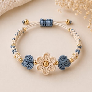 Peut inclure: Un bracelet en macramé avec une fleur centrale crème au cœur doré, entourée de deux nœuds en forme de feuilles bleues. Le bracelet est composé de cordons tressés alternant le crème et le bleu, agrémentés de petites perles dorées et d'un nœud coulissant.