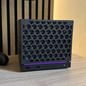 Könnte beinhalten: Ein würfelförmiges Computergehäuse in Mattschwarz mit einer markanten Frontblende im Wabenmuster. Das Gerät verfügt über einen dünnen, horizontalen violetten LED-Lichtstreifen, zwei USB-Anschlüsse, einen kleinen Schlitz und einen runden Power-Knopf.