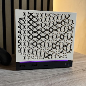 Könnte beinhalten: Ein rechteckiges Computergehäuse mit einer weißen Frontplatte in einem geometrischen, sternförmigen Gittermuster. Die Basis ist schwarz und verfügt über zwei USB-Anschlüsse, eine kleine Kontrollleuchte und einen runden Einschaltknopf, akzentuiert durch einen dünnen violetten LED-Lichtstreifen.