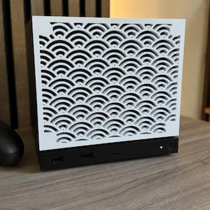 Könnte beinhalten: Ein rechteckiges Computergehäuse mit einer weißen Frontplatte, die ein sich wiederholendes, halbkreisförmiges Wellenmuster aufweist. Die Basis ist schwarz und enthält zwei USB-Anschlüsse, einen runden Power-Knopf mit weißem Licht und einen Schlitz.