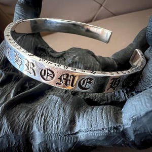 May include: A polished silver cuff bracelet featuring an open design and a flat band. The exterior surface is engraved with the text CHROME HEARTS in a bold, gothic-style font, interspersed with small cross symbols along the entire length of the band.