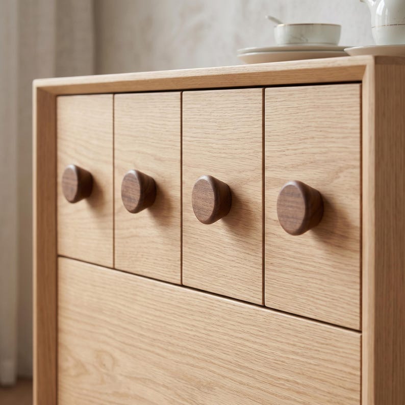 Puede incluir: Una c&oacute;moda de madera clara con cuatro cajones superiores verticales, cada uno equipado con un peque&ntilde;o pomo redondo de madera de color marr&oacute;n oscuro. La veta de la madera clara es visible en los paneles frontales y el marco del mueble.