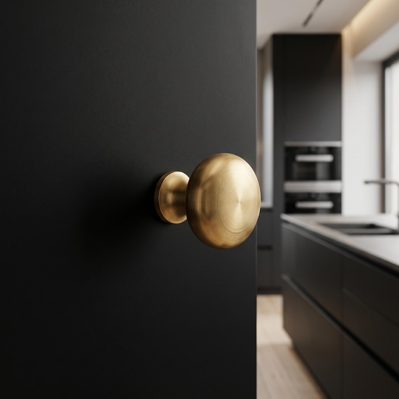 May include: A circular, brushed brass door knob with a smooth, rounded face and a cylindrical base, mounted onto a solid matte black door surface. The metal finish displays a subtle radial texture, reflecting light across its curved surface.