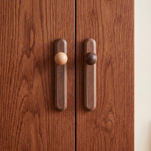 Puede incluir: Dos tiradores de armario verticales de madera montados en una puerta de madera de color marr&oacute;n rojizo con veteado. Cada tirador tiene una base rectangular larga con extremos redondeados y un pomo circular. Un pomo es beige claro y el otro marr&oacute;n.