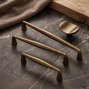 May include: A collection of antique brass hardware including two long, slender cabinet pulls with tapered ends and one small, rounded, concave cabinet knob. The metal has a brushed, weathered finish with visible dark highlights and a smooth texture.