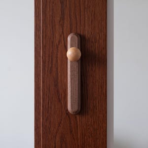 Puede incluir: Un tirador de puerta de madera marr&oacute;n oscuro que presenta una base rectangular vertical con extremos redondeados y un pomo esf&eacute;rico liso de color claro fijado en el centro. El tirador est&aacute; montado sobre una superficie de madera veteada.