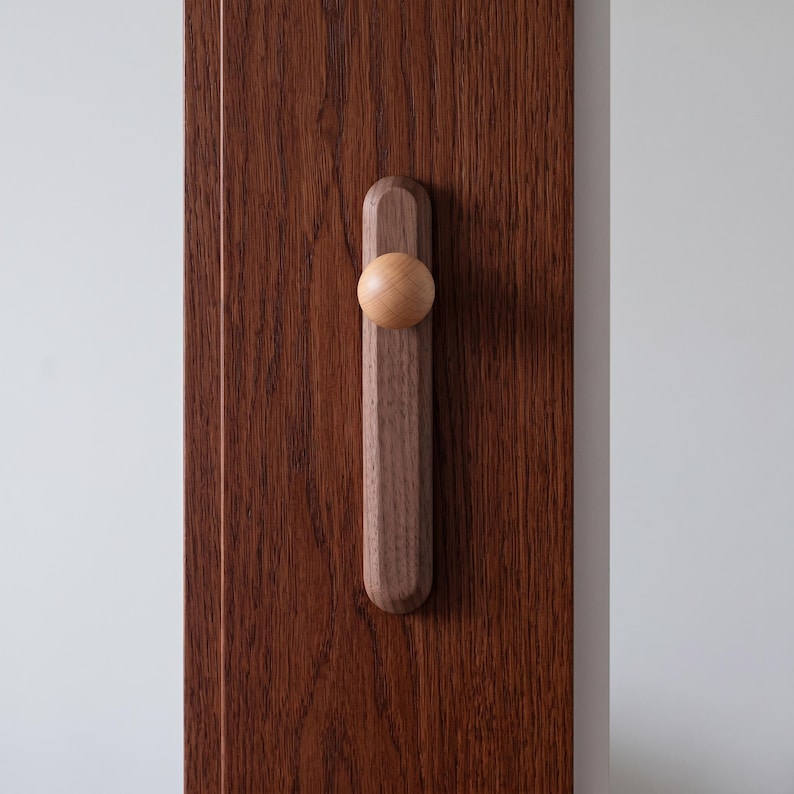 Puede incluir: Un tirador de puerta de madera marr&oacute;n oscuro que presenta una base rectangular vertical con extremos redondeados y un pomo esf&eacute;rico liso de color claro fijado en el centro. El tirador est&aacute; montado sobre una superficie de madera veteada.
