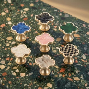 May include: Seven clover-shaped cabinet knobs with brass-toned metal bases. The tops feature various patterns and colours, including solid blue, dark grey marble, green, light pink, cream, grey marble, and a black and tan checkered design.