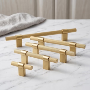 Peut inclure: Une collection de quatre poignées de placard dorées de longueurs variées, chacune composée d'une barre cylindrique à la surface texturée et moletée, avec deux supports de montage rectangulaires lisses. Les poignées sont disposées en quinconce.
