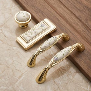May include: A collection of gold-toned cabinet hardware including one round knob, one rectangular pull, and two arched handles. Each piece features a central cream-colored ceramic inlay decorated with a delicate green and purple floral vine pattern.