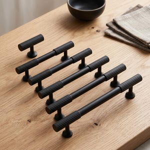 May include: A collection of six matte black cabinet pulls of varying lengths, each featuring a cylindrical bar with a textured, ribbed grip section on both ends and smooth, circular mounting posts at the base for attachment to furniture.