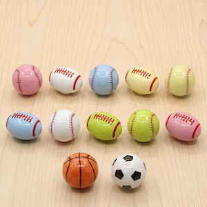 Op de afbeelding: Een collectie van twaalf keramische kastknoppen in de vorm van sportballen, waaronder honkballen, footballs, een basketbal en een voetbal. De knoppen zijn uitgevoerd in diverse kleuren zoals roze, wit, blauw, geel en groen met geschilderde details.