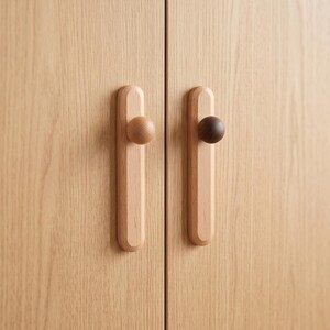 May include: Two light-coloured wooden cabinet doors featuring vertical rectangular backplates with rounded ends. Each door has a circular knob attached to the centre of the backplate, with one knob in a light wood tone and the other in a dark wood tone.