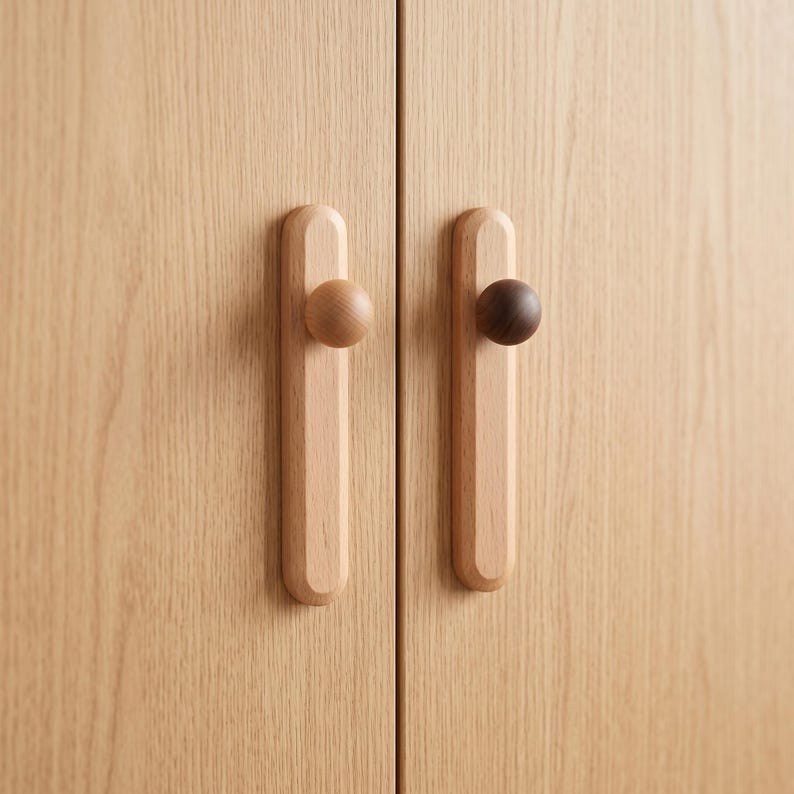 May include: Two light-coloured wooden cabinet doors featuring vertical rectangular backplates with rounded ends. Each door has a circular knob attached to the centre of the backplate, with one knob in a light wood tone and the other in a dark wood tone.